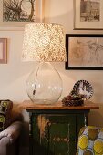 Lamp with clear glass balloon base and small Christmas arrangement of fir cones on vintage cabinet below pictures on wall