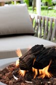 Moderner Aussengrill mit brennenden Holzscheiten vor Polsterstuhl