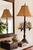 Old wooden jewellery box and silver urn on surface with ornate brass lamp stand. A rectangular wooden framed mirror hangs on the wall