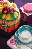 Crocheted tea cosy next to teacup with gilt pattern and tea caddy