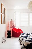 Red upholstered armchair next to stylised tree used as clothes rack in corner of white bedroom with white interior window shutters