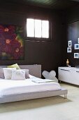 Modern double bed against black-painted wall with floral painting next to transom window in corner of room