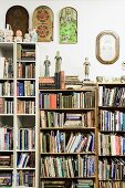 Figuren auf Büchern verschieden hoher Bücherregalen an Wand mit gehängtem Wandschmuck