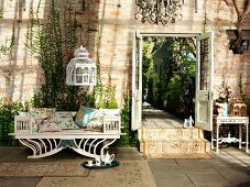 Rustikales Ambiente mit antiker Sitzbank und von Decke abgehängter Vogelkäfig neben offener Flügeltür mit Gartenblick