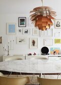 Marbled dining table and shell chairs in front of white sideboard; collection of framed pictures on wall and designer lamp hanging from ceiling above dining table
