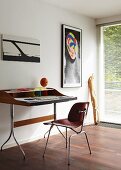 Moderner Schreibtisch mit Stuhl vor weißer Wand mit abstrakten Bildern, im Hintergrund Fenstertür mit Jalousie, davor lehnt ein Kunstobjekt aus Holz an der Wand