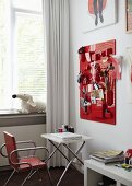 Teenage girl's bedroom with various utensils in containers on red organiser board above laptop on white folding table and metal, upholstered chair in front of window with white curtain
