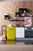 Electric kitchen appliances and white kitchen base unit in a row below various items of crockery on black shelves on brick wall