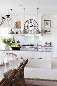 weiße offene Küche mit Pendelleuchten und Wanduhr, rustikaler Esstisch mit Korbstühlen