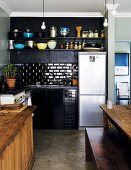 Corner of simple, black kitchen with stainless steel fridge-freezer and partially visible wooden table and bench in traditional setting