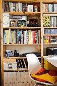 Volle Regalwand in Arbeitszimmer mit weißem Schalenstuhl und weißem Schreibtisch; auf dem Stuhl ein buntes Kissen
