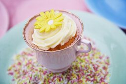 A cupcake decorated with light frosting and a marzipan flower in a cup