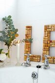 Lit candle, vases of flowers and large letters hand-crafted from corks leaning on white tiled wall on edge of bathtub