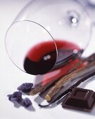 A glass of red wine lying on its side, with candied violets, dark chocolate, a vanilla pod and liquorice roots