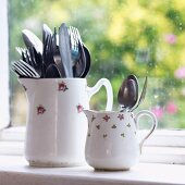 Large and small items of cutlery in two old china jugs with floral patterns in front of window