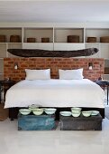 Doppelbett mit weisser Bettwäsche vor halbhoher Ziegelwand und Vintage Schüsseln auf Truhen am Bettende im Schlafzimmer