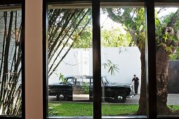View through window of black, classic saloon car and person dressed in black in front of high, white wall