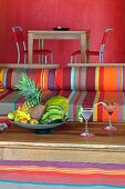 Sitzplatz mit bunt gestreiften Polstern und Schale mit tropischen Früchten auf Holztisch; Esstisch vor rot getönter Wand im Hintergrund