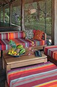 Fröhlicher Sitzplatz mit bunt gestreiften Polstern und Schale mit tropischen Früchten auf naturbelassenem Holztisch