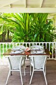White rattan chairs around table with place settings on wooden floor of veranda with tropical, bushy palm in background