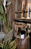 Still-life arrangement of cushions and decorative plants next to tree trunk table and bottle in front of glasses on wooden stool