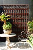 Palm-like plant on stone table and wire chair on patio with ornate metal grid on dark wall