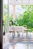 View through an open door onto a terrace with checkerboard tiling and filigree wrought iron chairs at a table set in white