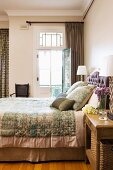 Double bed with elegant bedspread and matching scatter cushions next to open balcony door in traditional, elegant bedroom