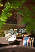 Bowl next to cushion on wooden bench and hand-made miniature sailing boot in pool