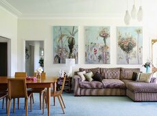 Dining area with wooden retro chairs and couch against wall below floral paintings in living room with stucco frieze running around edge of ceiling