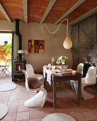 Simple wooden table and white plastic shell chairs below teardrop-shaped pendant lamp in simple country house with ribbed ceiling