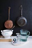 Jug of water, glasses and soup tureen in front of vintage pans hanging on black wall