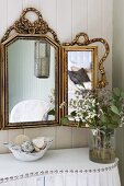 Glass vase of flowers on curved console table with white cover and decorative studs below gilt-framed, triple mirror on wooden wall