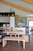 Pale wooden bench and stools at dining table in open-plan, modern kitchen with wood-beamed ceiling