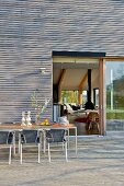 Tisch und Stühle auf Terrasse vor zeitgenössischem Haus mit Holzverschalung und Blick durch offene Terrassentür ins Wohnzimmer