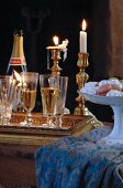 Gefüllte Sektgläser auf Tablett vor Messing-Kerzenständern mit brennenden Kerzen und Porzellanschale mit Konfekt auf Tischtuch