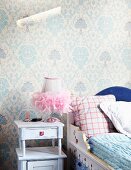 Painted wooden child's bed against wallpaper with traditional pattern and next to lamp with tulle lampshade on romantic vintage bedside table