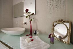 Toiletries on designer washstand with counter-top basin on glass counter and flower in vase next to vintage mirror