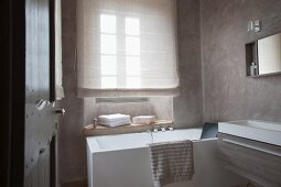 Designer bathroom - view through open door of bathtub below window with drawn blind and modern trough-style wash basin against wall