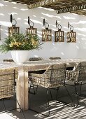 Pattern of light and shade falling through pergola onto rattan chairs with metal frames at rustic wooden table; row of lanterns hanging on wall