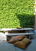 Ethnic cushions on floor in front of rustic wooden bench and table in sunny courtyard with creeper-covered wall