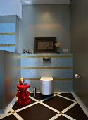 Wandtoilette in elegantem Badezimmer mit pastellblauen und goldenen Mosaikfliesen; im Vordergrund ein rotes Beistelltischchen mit einem goldenen Dekoobjekt