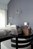 Sessel mit Holz Rückenlehne und naturfarbenem Polsterbezug vor Doppelbett unter Hängeleuchten mit Glasschirm vor grauer Wand