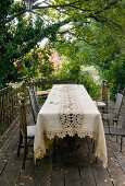 Tablecloth with lace insert and metal chairs on wooden terrace in garden with dense vegetation
