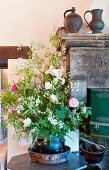 Large bouquet in ceramic pot on table in front of old metal jugs on stone mantelpiece of partially visible fireplace