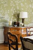 Baroque chest of drawers against floral wallpaper and artistically crafted dining chairs
