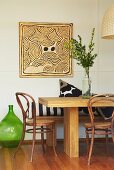 Square, expressionist artwork on wall; solid wooden table, bench, vintage bistro chairs and old oil bottle used as floor vase in foreground