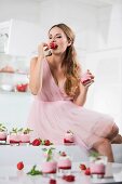Germany, Young woman biting strawberry, glasses with strawberry yogurt