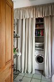 Blick durch offene Tür auf eine Waschmaschine unter Regalböden hinter bodenlangem Vorhang