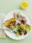 A slice of spinach quiche on a plate with ham and salad